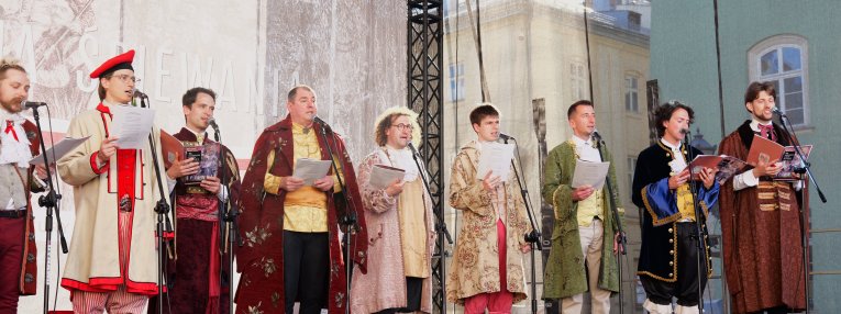 Grupa mężczyzn w historycznych strojach śpiewa na scenie plenerowej, stojąc przy mikrofonach i trzymając teksty w dłoniach.